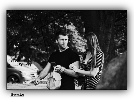 A black-and-white street photograph capturing a young couple walking through a park. In the background, trees line the scene and a parking area filled with cars sits just beyond the park — a typical urban setting wrapped in subtle contrasts of light and shadow.

The man is dressed in a black t-shirt and dark trousers, carrying both a small backpack (likely his partner’s) and a smartphone in the same hand. His posture is relaxed, yet attentive — his gaze fixed on the woman with a look of intrigue, perhaps even tenderness. His hair is short, his appearance neat, and his expression quietly expressive.

The woman walks beside him in a knee-length polka dot dress. She’s gently holding onto his arm, leaning in slightly as if explaining something with intent or sharing a meaningful thought. Her face shows focus, and her hairstyle is smooth and natural.

The image conveys an intimate, quiet moment between two people — suspended in the hum of everyday city life, in a gesture, a glance, and silent understanding.