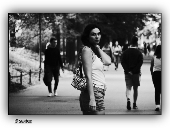 A black-and-white street photograph taken in a park shows a wide alley where many people are walking — blurred silhouettes of passersby appear in the background, alongside trees that frame the path and build a calm urban landscape.  
In the foreground, a woman has seemingly stopped and turned toward the photographer. Her gaze is intense, almost surprised — wide-open eyes draw attention, creating a sense of spontaneous connection. She’s dressed in a sleeveless top and shorts, a light, summery outfit suited to warm weather. Over her shoulder hangs a distinctive zebra-patterned bag — a bold accent in black and white. One of her hands is raised, likely reaching toward her neck or nape — as if brushing back her hair or reacting to the moment.  
The composition captures a fleeting instant and an accidental meeting of gazes — classic hallmarks of street photography, where everyday life gains drama and intimacy.