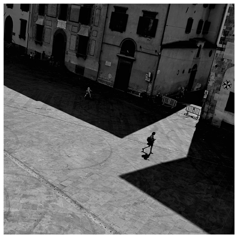 A black and white photo from higher up of an Italian square with historic buildings surrounding it. A single person walks across the sun-lit part of the stone-paved square, casting a shadow. 