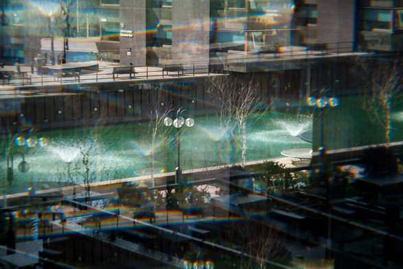 Photo of a rectangular concrete pool filled with green water, taken from a above and to one side. The image is optically split into five copies lined up such that the near side of the pool repeats alond the line of its nearest bank, in the water are low wide fountains, along the bank are streetlamps with clusters of four round lights and in the foreground is a shadowy balcony, all repeated and overlapping with themselves.