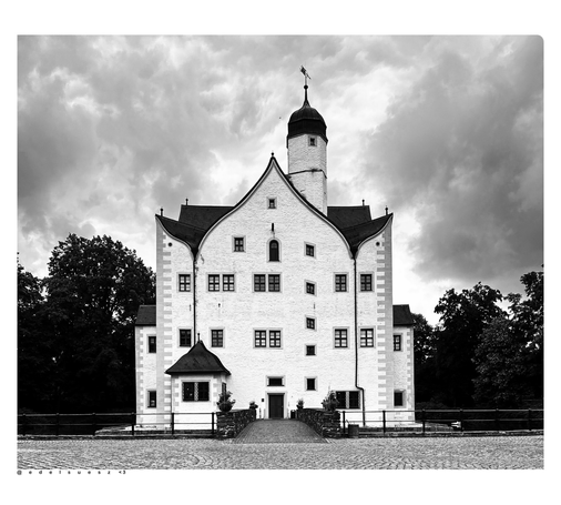 Schwarzweißfotografie: Seitenansicht vom Wasserschloss, aus der Zeit der Renaissance, heller Putz, kastenförmiger Bau mit kleinen Fenstern und geschwungenen Giebelformen, plus außermittigen Turm, gepflasterter Platz davor, links und rechts dunkle Bäume, grauer wolkiger Himmel darüber 