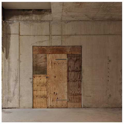 A photo of an unfinished concrete interior space with exposed walls and ceiling showing construction joints and some water staining. In the center sits a makeshift wooden door constructed from plywood panels with visible wood grain, metal hinges and a padlock.