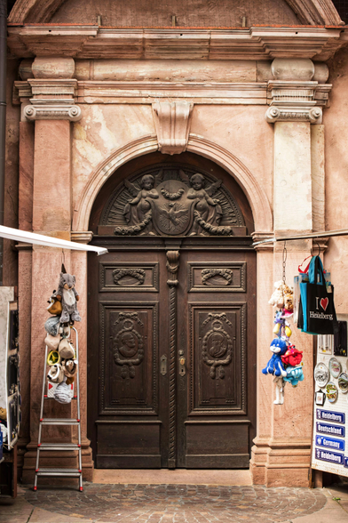 Säulenbewehrte, pompöse, zweiflügelige Nebeneingangstüre einer Kirche. Auf den Türflügeln ist jeweils ein Konterfei eines bärtigen Mannes geschnitzt. Im oberen Teil der in einem Rundbogen sitzt sind zwei nackte Kinder mit Flügeln eingearbeitet. Rechts und links von der Türe hängt Tand von den ausstehenden Klappen der Andenkenverkäuferwagen.