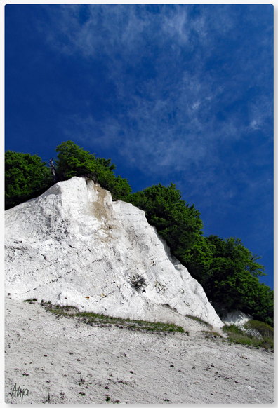 Das Bild zeigt einen Steilhang an einer Küste, der Blick geht vom Ufer aus nach oben: weißer Kreidefelsen, eine grüne Baumreihe und ein - fast - strahlend dunkelblauer Himmel darüber.

The picture shows a cliff on a coast, the view from the shore looking upwards: white chalk cliffs, a row of green trees and an - almost - brilliant dark blue sky above.