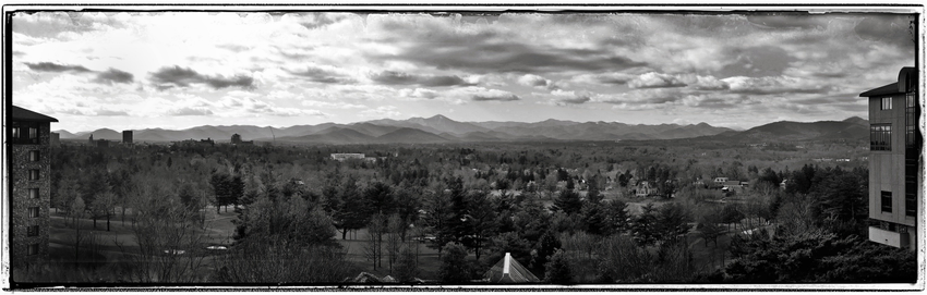A monochrome picture of the countryside full of trees... and, in the distance, mountains. The sky is peppered with storm clouds and the glare of the afternoon sun.