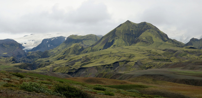 Photograph of smooth and brown hills and small mountains covered with lime green moss carpet. A glacier is hidden in fog and clouds behind. Bushes and red and short grass can be seen in the foreground at the bottom of the picture.