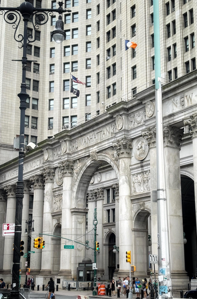 Closeup of the facade of the Manhattan Municipal Building as seen from Centre Street. A colonnade of Corinthian columns looms over tiny pedestrians, traffic signals, and orange traffic barriers. In between the central columns are three arches, the middle arch nearly twice as tall as the side ones. Bas-relief panels are sculpted above the side arches. Atop the columns is a frieze displaying the words NEW AMSTERDAM, MANHATTAN, and NEW YORK (with YORK out of frame past the right edge) in giant Roman font alongside Roman numerals. Behind the colonnade, the main facade of the building with its grid of office windows rises past the top of the photo. Two lampposts, a black-painted old-fashioned one and a more modern one, stand at left and right in the foreground, framing the scene.