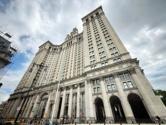 A wider-angle view of the facade of the Manhattan Municipal Building as seen from Centre Street. Here the entire edifice can be seen, distorted by the lens as it rises and recedes from camera into a cloudy sky. From this angle, the gilded statue “Civic Fame” can be glimpsed at the apex of the central tower. The fact that the building has some significant amount of space around it, and is set apart from other buildings in the area, emphasizes its massive size.