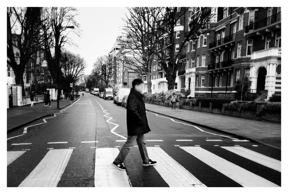 Black and white photo of me in a dark winter coat, jeans, and sneakers crossing Abbey Road in London, in homage to the Beatles album cover. I’m mid-stride and facing right, walking on a zebra crossing with wide white stripes. Painted zigzag lines mark the curbsides and the division between the two lanes of the road. Apartment buildings and leafless trees line the sidewalks under a featureless winter sky.