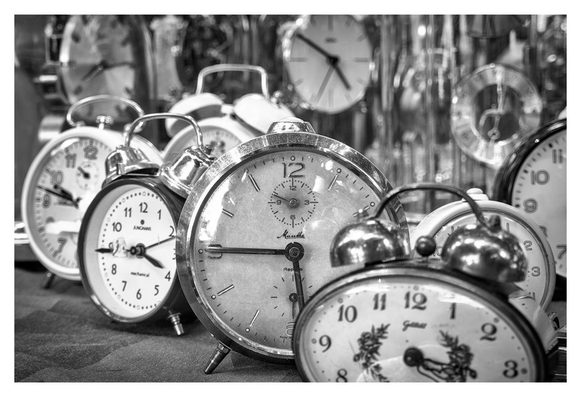The image is a black-and-white photograph featuring a collection of vintage analog alarm clocks. The clocks are arranged closely together, with some in sharp focus in the foreground and others slightly blurred in the background, creating a sense of depth. Some of the clocks have metallic bells on top, typical of traditional alarm clocks, and some have decorative elements or brand names visible on the dials. The hands of the clocks point to different times. The background is filled with more clocks, adding to the cluttered yet nostalgic atmosphere. The overall tone is monochromatic, emphasizing the textures and details of the clocks' surfaces.