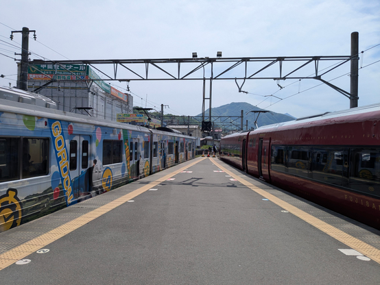 An einem Bahnsteig warten  Züge auf Gäste.
Links ein bunt gehaltener Bummelzug, rechts der tiefrote Express. 
