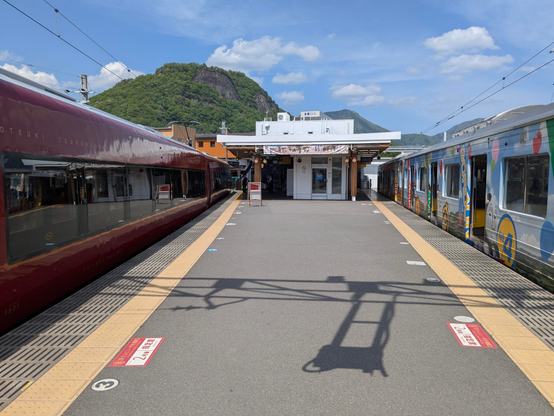 An einem Bahnsteig warten  Züge auf Gäste.
Rechts ein bunt gehaltener Bummelzug, links der tiefrote Express. 
Außerdem noch in der Mitte ein Bahnhofsgebäude. Im Hintergrund ein begrünter Hügel.
Im Vordergrund Schattenwurf der Oberleitung. 