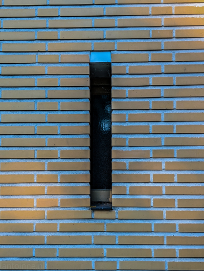 A brick wall, part of the exterior of Hilversum Town Hall. The bricks are a pale, yellowish-beige color with a smooth, matte finish. The most prominent feature of the image is a narrow, vertical window in the middle of the wall. The interior of the window is dark, but a faint, rippled texture can be seen on the glass.