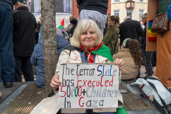A protest scene on Grand Parade, Cork, showing a person sitting on the ground among a crowd, holding a handmade sign that reads: "The terror State has Executed 50,000+ Children In Gaza." The person is draped in a flag of Palestine, and other demonstrators with placards and flags are visible in the background.