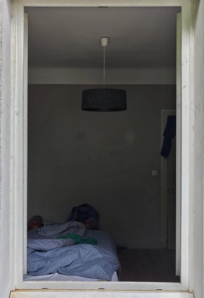 A view through an open window into a sparse bedroom with white walls and ceiling. One can see an unmade bed with blue bedding and dark pendant light fixture hanging above. The room appears minimally furnished.