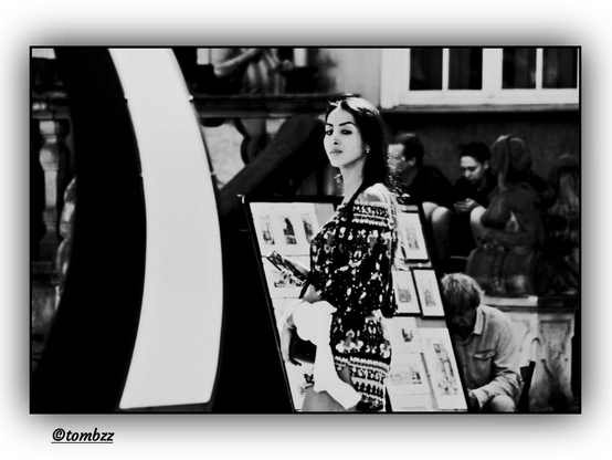 A black-and-white street photograph captures a young woman standing in front of an outdoor display of framed pictures arranged on a stand. She has long, loosely flowing hair and is wearing a patterned, lightweight summer dress that suggests a warm day and adds a subtle elegance to her silhouette. Her gaze is directed straight at the camera — she has clearly noticed the photographer, lending the image a sense of spontaneity and authenticity.
In the background, we see an urban space, likely a market or square, with several people visible — some seated, others strolling by. The framed artworks on display introduce an element of culture and creativity to the everyday scene. The overall composition evokes a nostalgic, quiet atmosphere, capturing a moment of city life in timeless monochrome.
