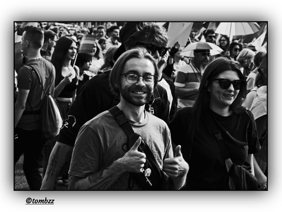 A black-and-white street photograph captures a crowd of participants at a pride march. In the center of the frame, in the foreground, stands a smiling couple — a woman and a man — clearly radiant, immersed in the celebratory atmosphere of the event. The man looks directly into the camera lens, establishing a warm connection with the photographer — his hands are raised in a thumbs-up gesture, full of positive energy and support.
 Their clothing is casual and comfortable, suited for a street demonstration — the man has short hair, wears a light T-shirt with a simple print, and carries a backpack slung over his shoulder. The woman stands beside him, also dressed in everyday attire — a light blouse, trousers, and sunglasses on her nose. Her face beams with a genuine smile and a sense of belonging.
 In the background, the crowd continues to march, accompanied by a waving rainbow flag — a symbol of equality and acceptance. Umbrellas in some participants' hands shield them from the intense sun, emphasizing the bright, summery climate of the march, which only adds to the joyful, festive atmosphere of this moment — one of solidarity and visibility for those who are often unseen.