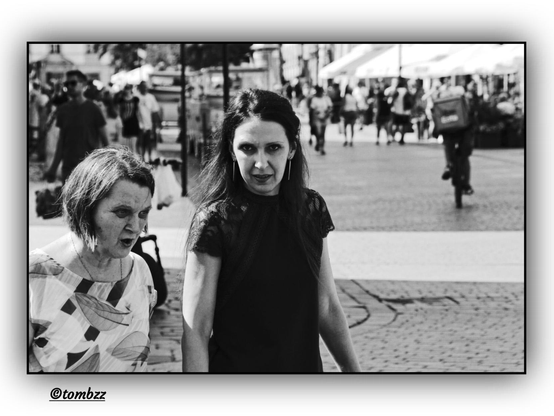 A black-and-white street photograph captures a moment in a lively city square. On a cobblestone street, two women stand at the center of the frame, deeply engaged in conversation. One of them, dressed in a simple dark top and wearing her hair loose, suddenly pauses and locks eyes with the camera — her intense gaze creates a striking moment of direct connection with the photographer, turning the image into a personal narrative.
 Her companion wears a boldly patterned blouse, adding visual contrast to the monochromatic palette. Both women appear absorbed in their shared moment, which is interrupted only briefly by the silent presence of the lens.
 In the background, pedestrians move through the scene and a cyclist rides off toward the right side of the frame. Further behind, café garden umbrellas line the edges of the market square, suggesting a relaxed and social atmosphere rather than the bustle of commerce. The composition pulses with urban life, yet the woman’s gaze halts that motion, capturing a fleeting second filled with meaning.