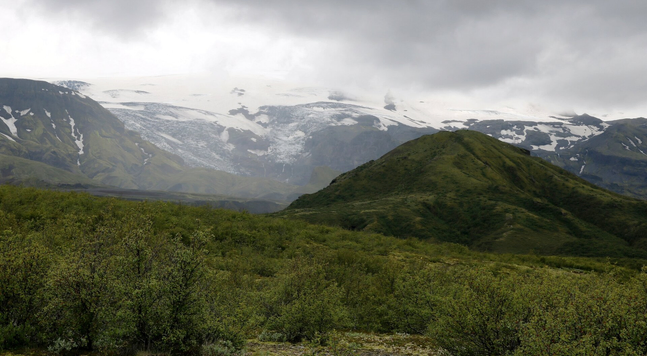 Photograph of a small conical mountain covered with green moss. Behind is a glacier, with dirty white snow over higher mountains, half hidden in fog and clouds. At the bottom, at the foot of the mountain, is a green slope with lush green bushes and small trees.