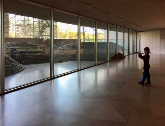April 2015 at Washington, DC National Gallery of Art in a ground or basement level room that has one side all windows. On the other side is an art installation of rounded mounds created by stacking thousands of flat pieces of rock. At one point a mound reaches the window and they’ve put stones on the inside of the window to give illusion that the mound has crept into building. A young boy in a plastic fire fighter help is standing a ways back from window holding an iPod to take a photo. 
The art installation is by Andy Goldsworthy, called Roof, it is from 2004-2005.

https://www.nga.gov/artists/28027-andy-goldsworthy