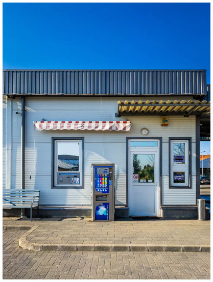 Gebäude, klein und einstöckig. Eingangs Tür und zwei Fenster sind hoch verspiegelt. Die Außenwand ist weiß, neben der Tür steht ein Automat, an dem man sich Lappen ziehen kann. Links im Bild eine weiße Bank, die angeschnitten ist.