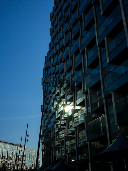 Photo looking up at the side of a very shiny tall building, all protruding glass balconies, a futurist cliff. The sun reflects from the midst of these, directly to the camera, caught in Jake Blues's biblical searchlight. At the bottom left is another class building in the distance, the rest of that side is clear blue sky.
