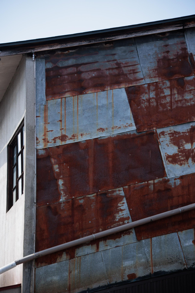 Rusty metal siding of a building in sunlight, featuring a mix of blue and reddish-brown hues, with a small window visible.