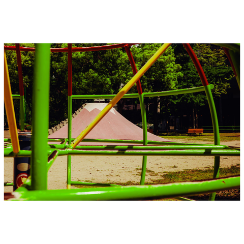 This photograph shows a public playground scene in Nagoya, with a bright green and orange jungle gym in the foreground framing a pink concrete play structure shaped like Mount Fuji in the distance. The Fuji structure is painted with a white peak to resemble snow and is lined with protruding black climbing grips on both sides. Trees fill the background, casting leafy shadows across the park.