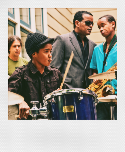 A snapshot from 2009. It shows a few members of a jazz band performing on a Fillmore Street sidewalk. In the foreground, a very young drummer, looking up and clenching his mouth as he observes the crowd in the distance. More importantly, in the background, a sharply dressed man in sunglasses leans in to a teenaged saxophonist and says something that the saxophonist appears to be considering.