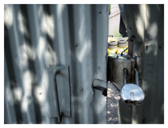 Photo of a double gate of gray corrugated metal, padlocked shut but with the right gate leaf bent and twisted, allowing for a glimpse into the sunlit yard beyond. Here we see two cars, one green and one yellow, parked with their fronts facing each other. Four tires are stacked two-by-two on the hood of the yellow car. The left gate leaf has a vertical bar handle. Dappled sunlight plays on the gate’s ridges.