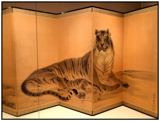 A folding screen of six silk panels, with patterned borders at top and bottom, displays a Japanese ink painting of a female tiger nursing her cub. The mother reclines against a sparse landscape while looking straight at the viewer with a warning glare, mouth slightly open. Her cub’s face is hidden behind her hind leg. The artist’s text and red seals can be seen vertically at the edge of the rightmost panel. The artwork seems to glow softly in tones of brown, yellow, and gold.