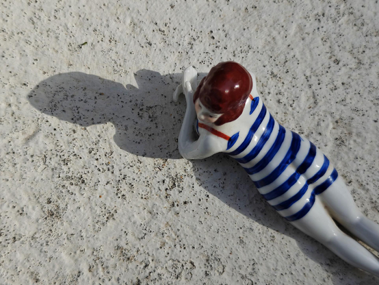 Top down photo of a ceramic figurine of a person in a blue and white striped bathing suit and a red cap lying on speckled stone surface, casting a sharp shadow in bright sunlight. The figure is lying on their belly and elbows, looking to the side.