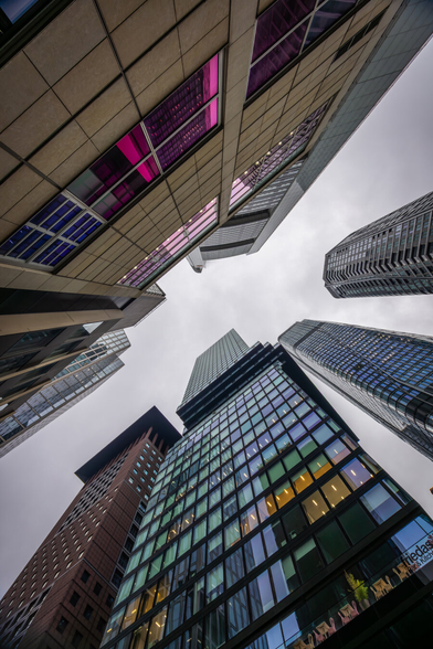 A dramatic upward view of modern skyscrapers in Frankfurt’s banking district. The buildings feature a mix of glass, steel, and concrete facades, with some windows reflecting pink and purple hues, others appearing blue and turqouise. The overcast sky adds contrast to the towering architecture converging toward the center of the frame.