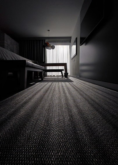 A modern hotel room with textured carpet flooring, a bed on the left, wooden desk and chair in the center, pendant lighting, and wall-mounted TV. Sunlight streams through the curtains covering window, creating shadows on the floor. The perspective and low angle of the camera create long shadow lines converging on the window in the distance.