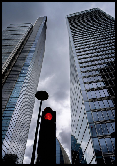 Dramatic low-angle view of modern glass skyscrapers towering overhead with a red traffic light in the foreground against a cloudy sky in London's financial district.