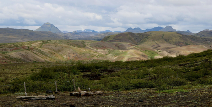 Photograph of colourful small and smooth hills, with hues of brown , beige, orange, red and grey, and some green from moss and small plants. Behind, peaks of higher mountains can be seen, under a cloudy sky. In front of the hills, in the bottom half of the picture, the brown ground is covered with green bushes and plants. A fence with a rope and three posts is visible at bottom left and piles of other posts.