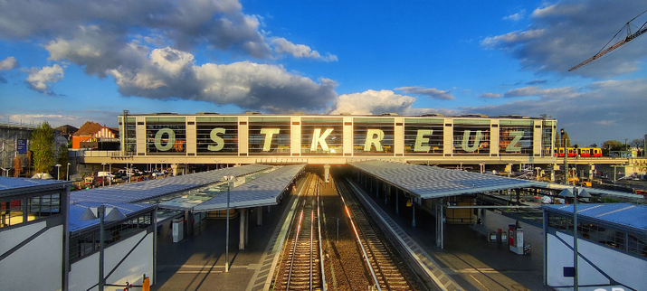 Der Bahnhof Berlin Ostkreuz wird von der Abend Sonne angestrahlt. Die Buchstaben des Bahnhöfe schimmern. Dahinter leicht bewolkter Himmel. Im Vordergrund die Gleise der S-Bahn
