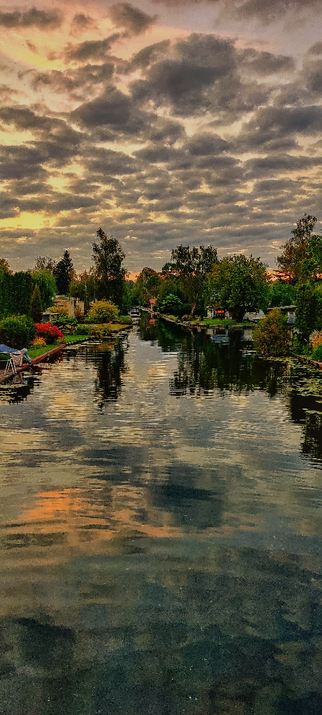 Zu sehen ist ein Wasserkanal in einem Wohngebiet. Der Himmel ist bewölkt und wird von der untergehenden Sonne diffus angestrahlt. Das ist ebenso in der Spiegelung im Wasser zu sehen. 
