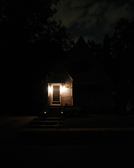Color photograph looking across a darkened street at night at the front of a house with bright lights shining on either side of the front door, the rest of the house set back into dark silhouette with the trees looming up beside and behind it under a partly cloudy night sky