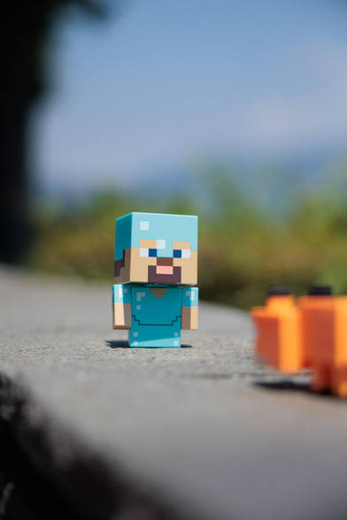 A Minecraft action figure of a character with blue attire stands on a stone surface, with blurred orange objects in the foreground.