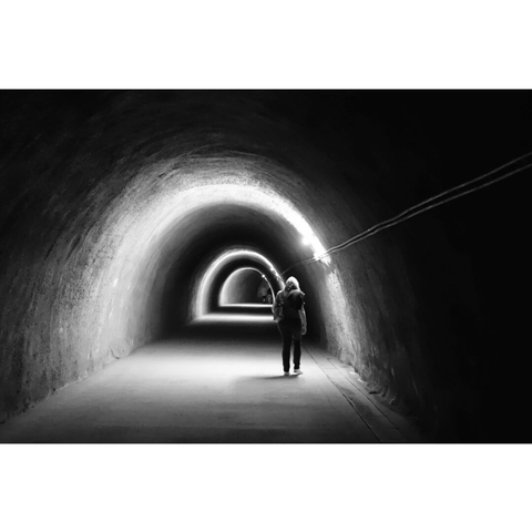 Black and white street photography taken in Luxembourg city. Somebody walks in the dark tunnel under the city. Half circles of light regularly illuminate the scene.