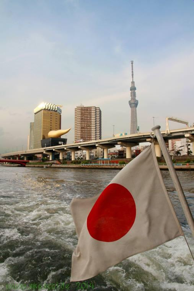 Perspektive vom Heck des Ausflugsschiffes auf den Fluß.
Im Vordergrund weht die japanische Flagge.
Im Hintergrund ist eine Hochstraße, die Gebäude der Asahi Bierbrauerei und der Skytree, ein Fernseh- sowie Aussichtsturm, zu sehen.