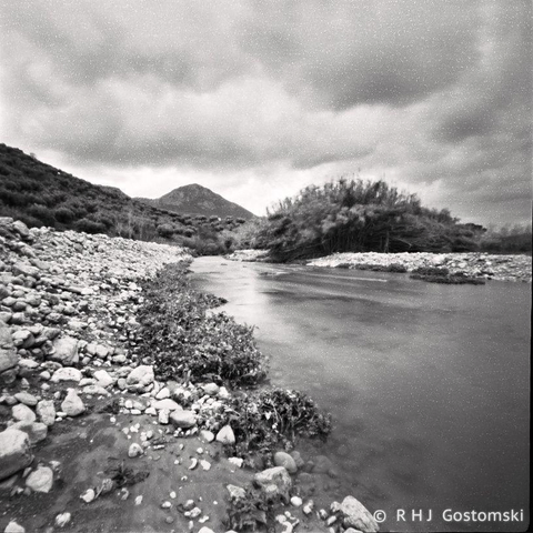 Pinhole black and white photo looking along a river with stones banks. Shrubs and a hill in the distance with a cloudy sky above. 