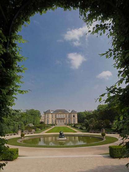 A classical French château viewed through an archway of green tree branches and leaves. The cream-colored stone building features symmetrical architecture with tall windows and a formal entrance. In the foreground, manicured gardens include a circular reflecting pool with a central sculpture, flanked by geometric hedges and pathways. The scene is set under a partly cloudy blue sky.