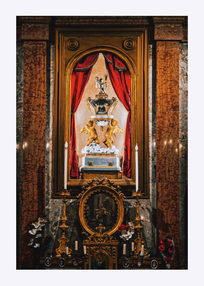 This is a photo of an ornate altar which is baroque in style. There are colours of gold and red, marble columns, a detailed religious statue containing a skull, golden lion and eagle and red velvet curtains. There two tall white candles on either side of this and there are intricate carvings and art underneath it.