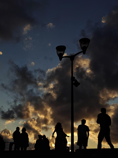As photo with silhouettes of several people waiting waiting for the sunset next to a tall street lamp against a twilight sky filled with high-contrast clouds. 