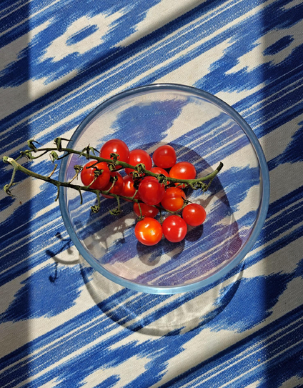 A bunch of fresh red cherry tomatoes on the vine sits in a clear glass bowl, placed on a blue and white striped table cloth with an abstract pattern. Bright sunshine makes for very vibrant colours.