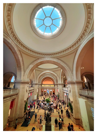 The Great Hall of the Metropolitan Museum of Art in New York, as seen from the second-floor balcony at the north end. A vast domed ceiling, with a central circular skylight ringed by lightbulbs, rests atop three massive arches. The middle arch leads into a second domed area with a similar arrangement of arches that in turn leads to a third. Each section of domes and arches recedes further from camera to create the impression of nested repeating geometric patterns. Beneath the side arches and the far middle arch are railed balconies overlooking the busy main floor, where people mill about the central information desk or the Egyptian statue at bottom frame. While the skylight is a luminous blue, the scene is predominantly in warm tones of tan and gold.