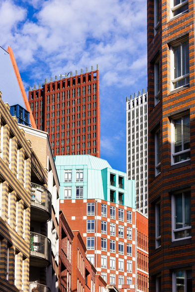 A colorful glimpse into the city center between two buildings with windows. Both buildings have horizontally striped walls. The left one is yellow and gray, and the right one is orange and brown. Further on, you see a low, horizontal, orange building, with a round green building above it. Above that, another tall, brown-red office building, and another one to the right, but white. Above it all, a beautiful blue sky with white clouds.