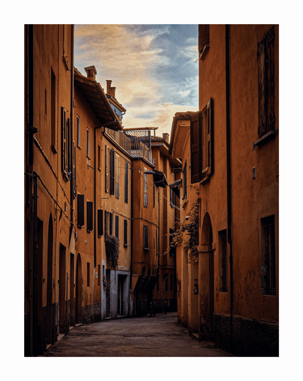 This is a photograph of a narrow street (Ca' Selvatica) in a rustic the Italian city of Bologna. The buildings are painted in warm terracotta and yellow hues and they eature wooden shutters, balconies, and a few potted plants. The sky above is partly cloudy with soft, golden sunlight which is casting long shadows.